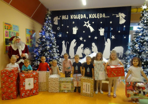 Uśmiechnięte dzieci trzymają prezenty, w tle figura Mikołaja.